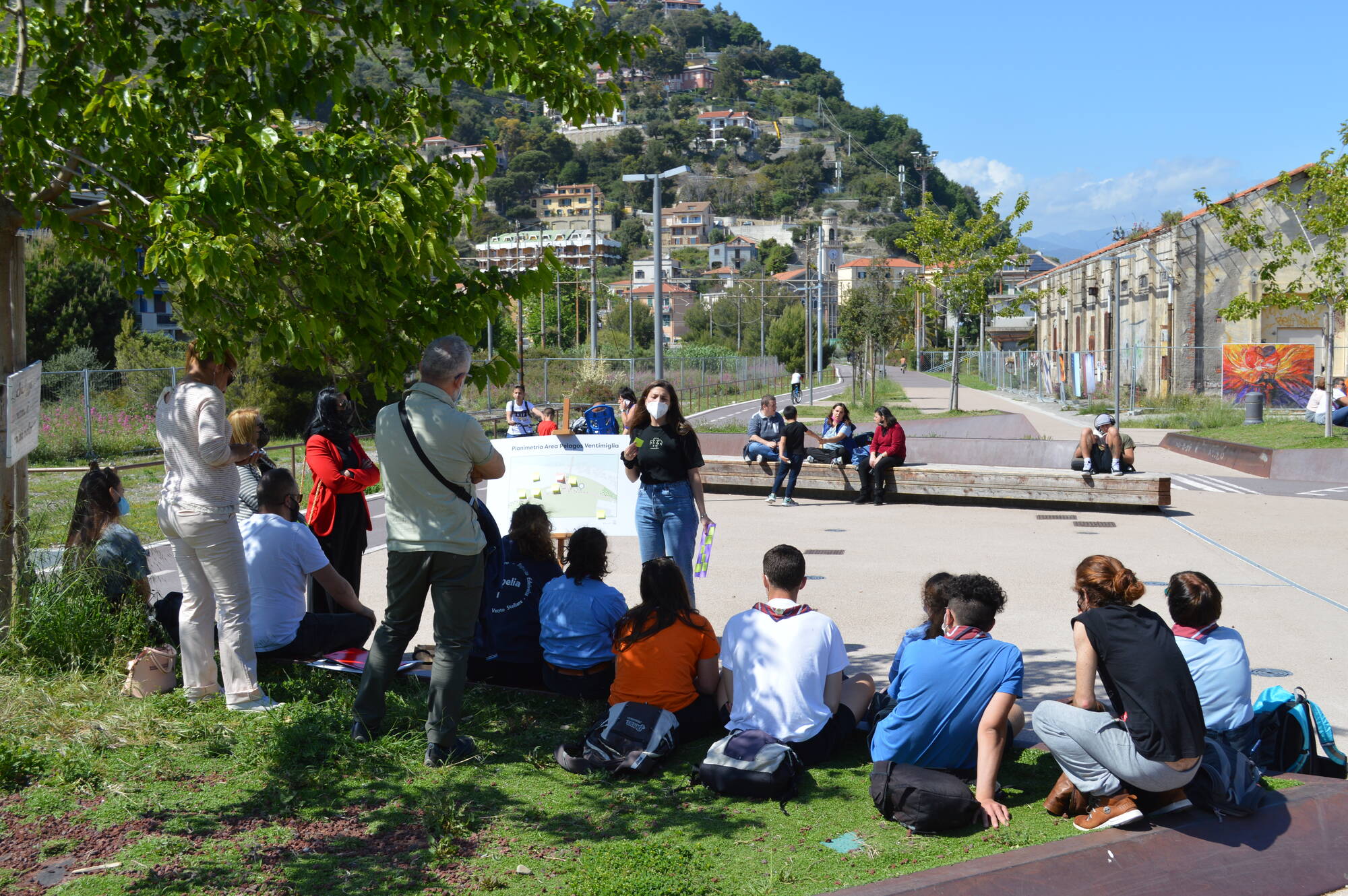 Partecipanti del progetto di co-design "Lo Spazio Pubblico" sono seduti all'aperto per ascoltare una presentazione. Sullo sfondo della fotografia la zona di Ventimiglia oggetto del progetto