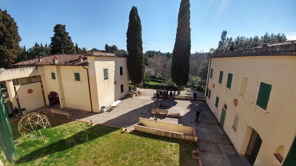 Cortile di una villa con prato e alberi, persone raccolte vicino a due cipressi sotto un cielo sereno.