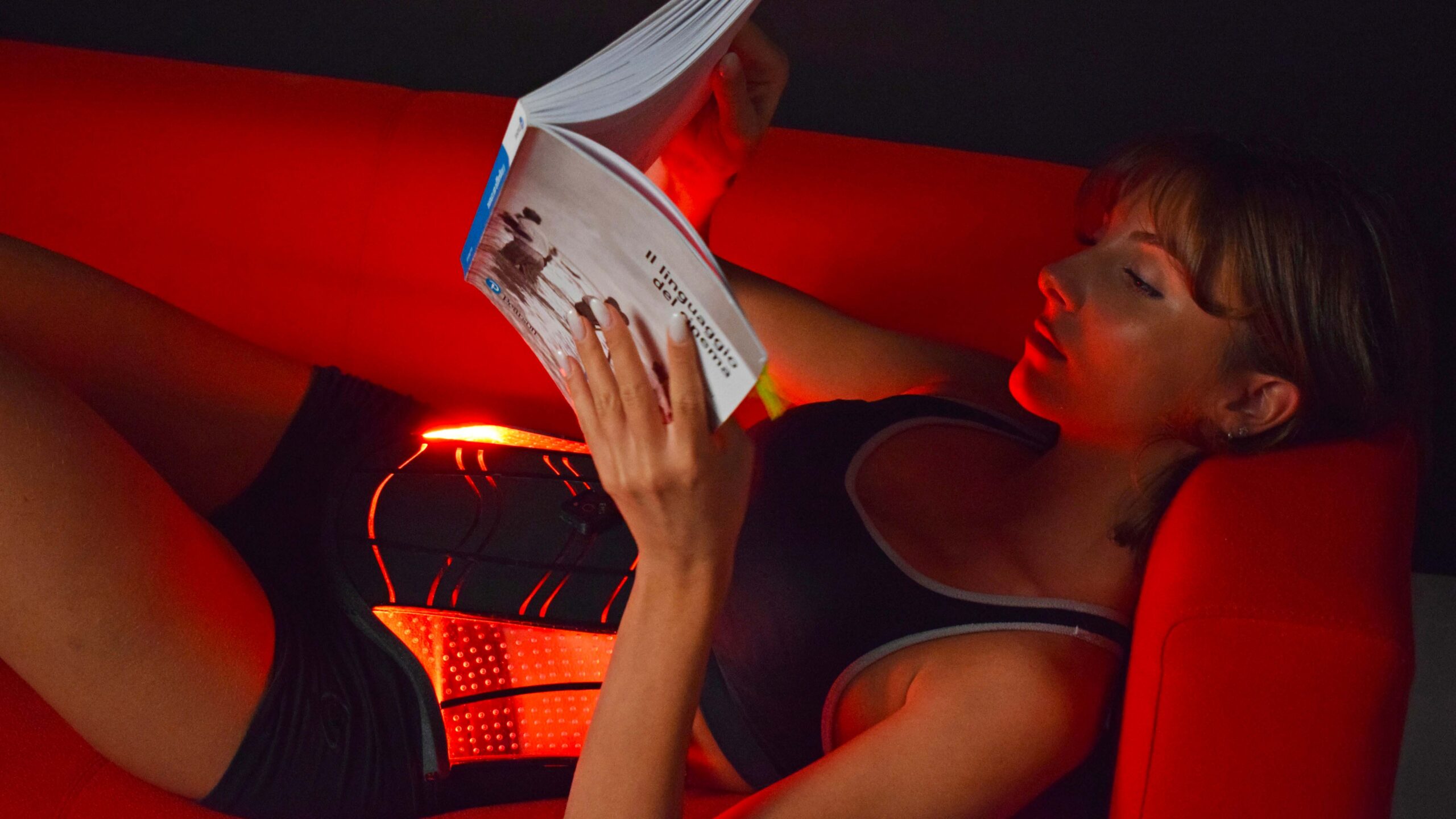 Donna reclinata su divano rosso, legge un libro con cintura massaggiante riscaldata illuminata mentre si rilassa.