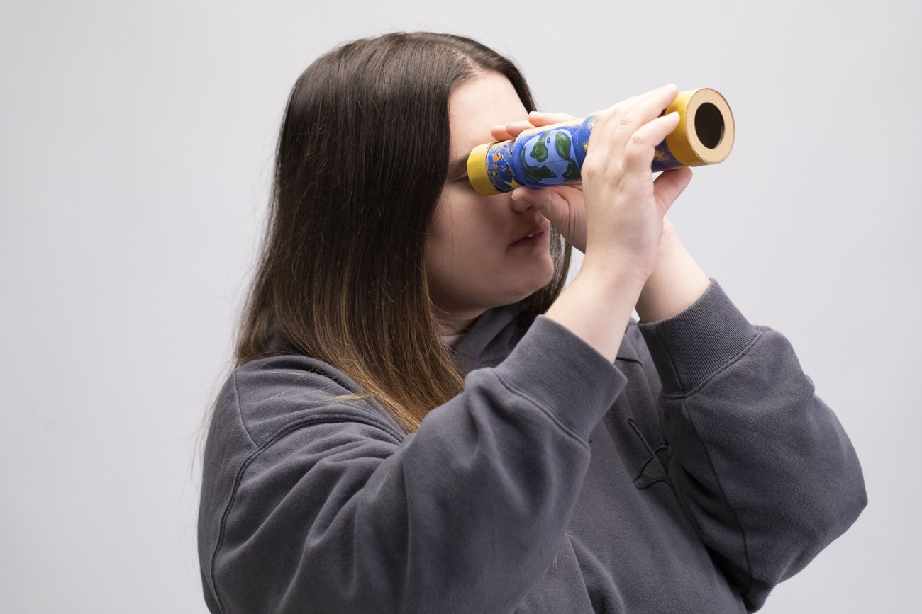 Donna guarda attraverso un caleidoscopio colorato su sfondo chiaro. Indossa una felpa grigia.