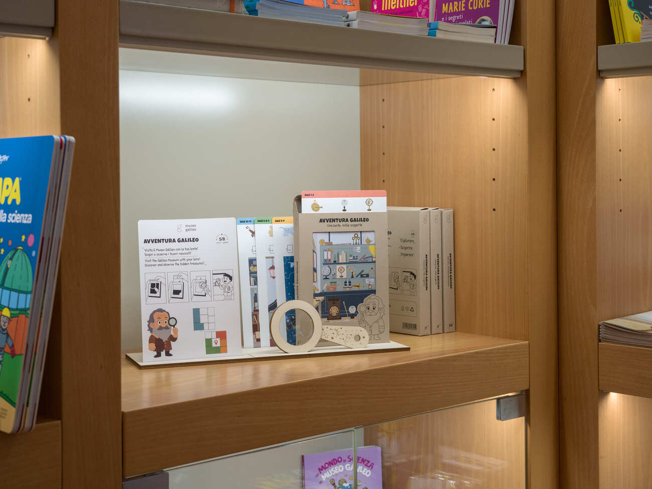Libri per bambini e giochi didattici Avventura Galileo esposti su una mensola in una libreria illuminata.