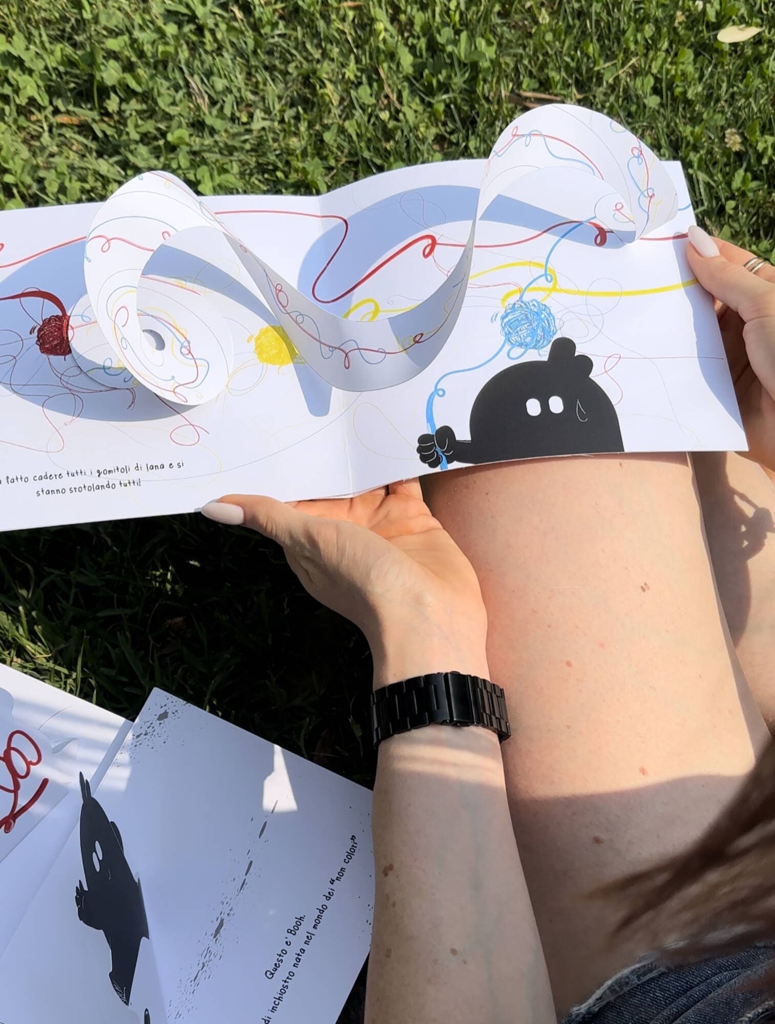 Persona all'aperto sfoglia un libro pop-up colorato, seduta sull'erba con un orologio nero al polso.