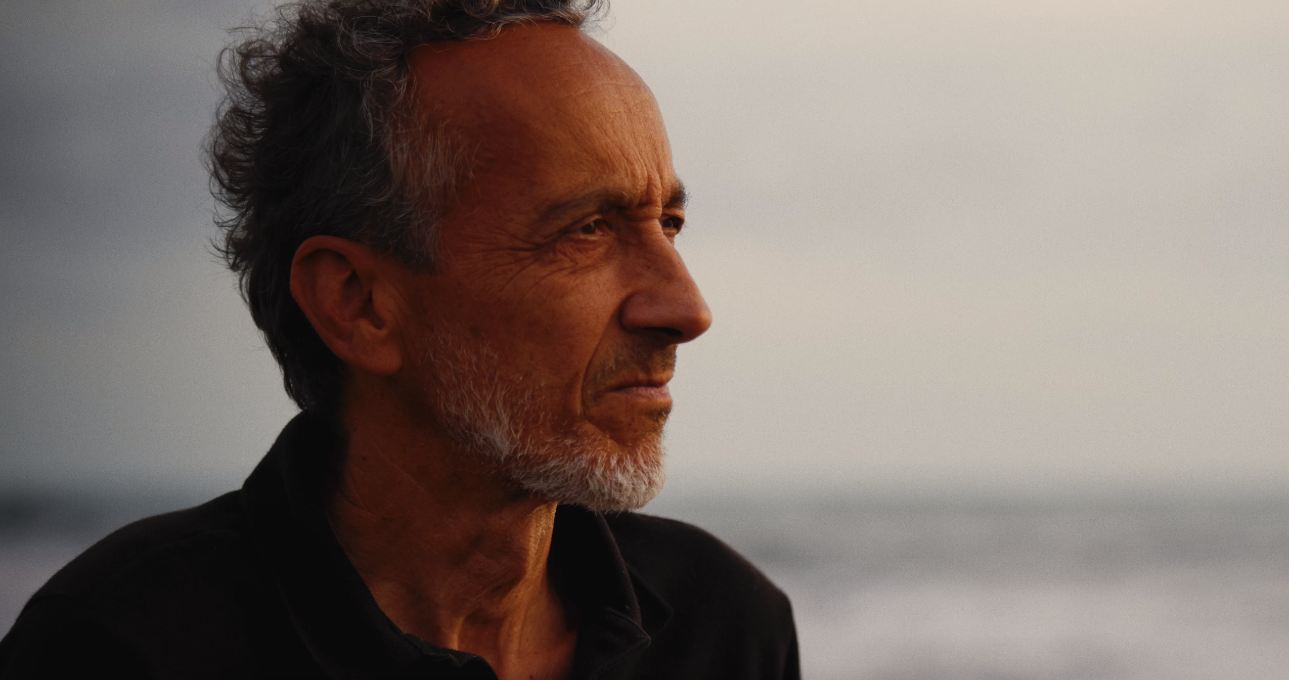 Ritratto di uomo maturo con barba grigia in controsole, sfondo del mare sfocato.