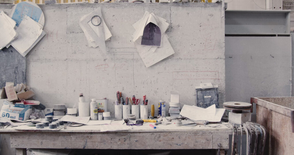 Banco da lavoro disordinato in officina con strumenti, documenti e materiali vari su sfondo di cemento.