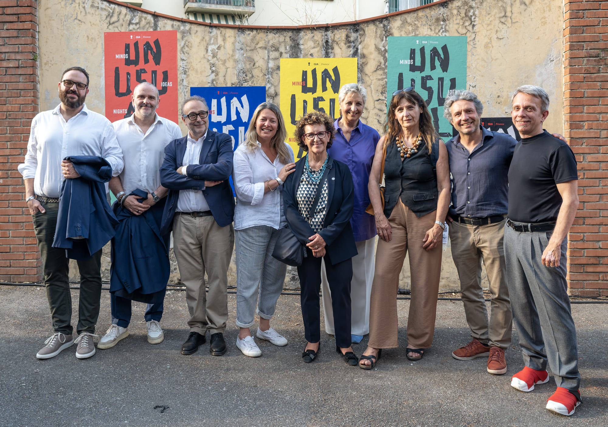 Gruppo di nove persone in posa davanti a manifesti colorati su un muro all'aperto, atmosfera amichevole e informale.