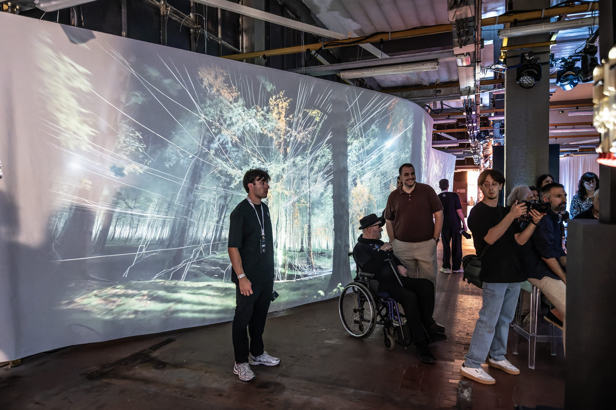 Persone in una mostra interattiva con proiezioni di foreste e luci, atmosfera immersiva e contemplativa.