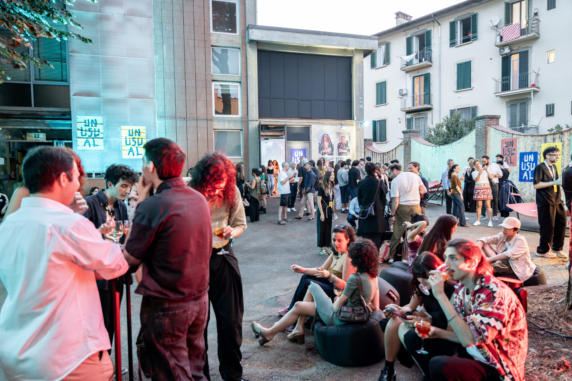 Persone socializzano a un evento all'aperto in città, illuminate da luci colorate, fra edifici residenziali e segnaletica UNUSUAL.