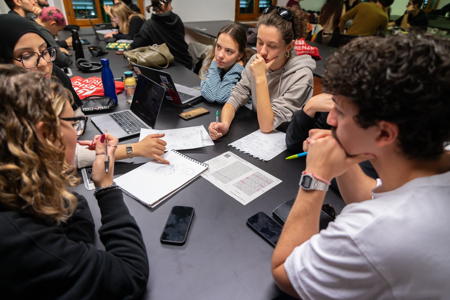 Studenti universitari collaborano su un progetto attorno a un tavolo, con laptop, telefoni e appunti.