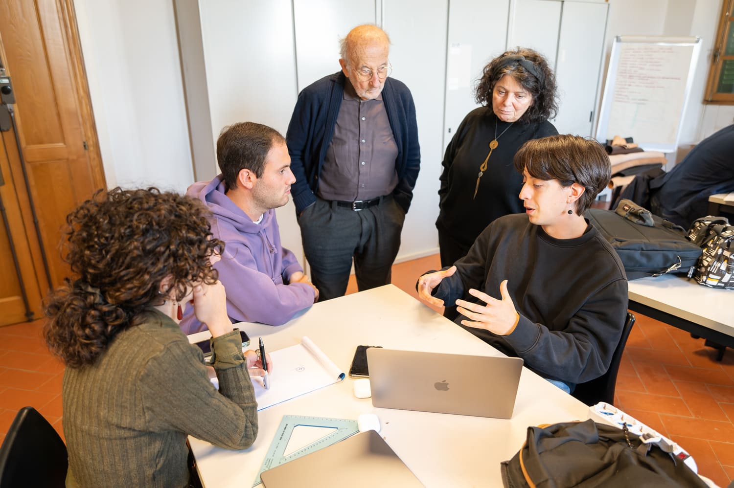 Gruppo di persone discute attorno a un tavolo con laptop, in un ambiente educativo collaborativo.
