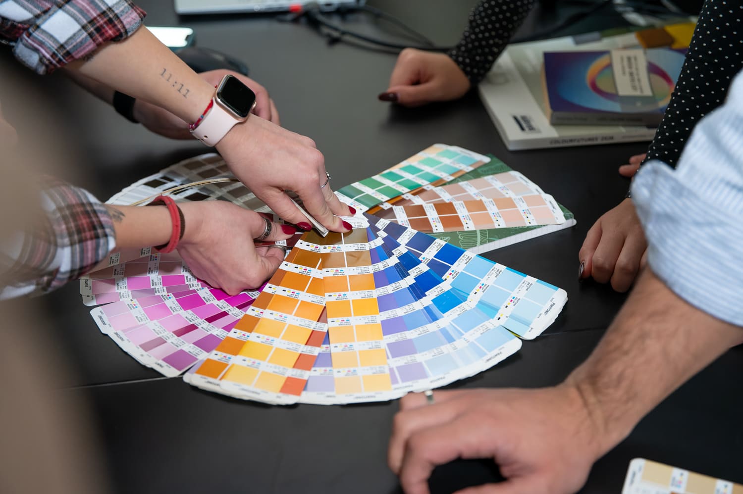 Persone selezionano colori da una mazzetta Pantone su un tavolo nero in un contesto creativo.