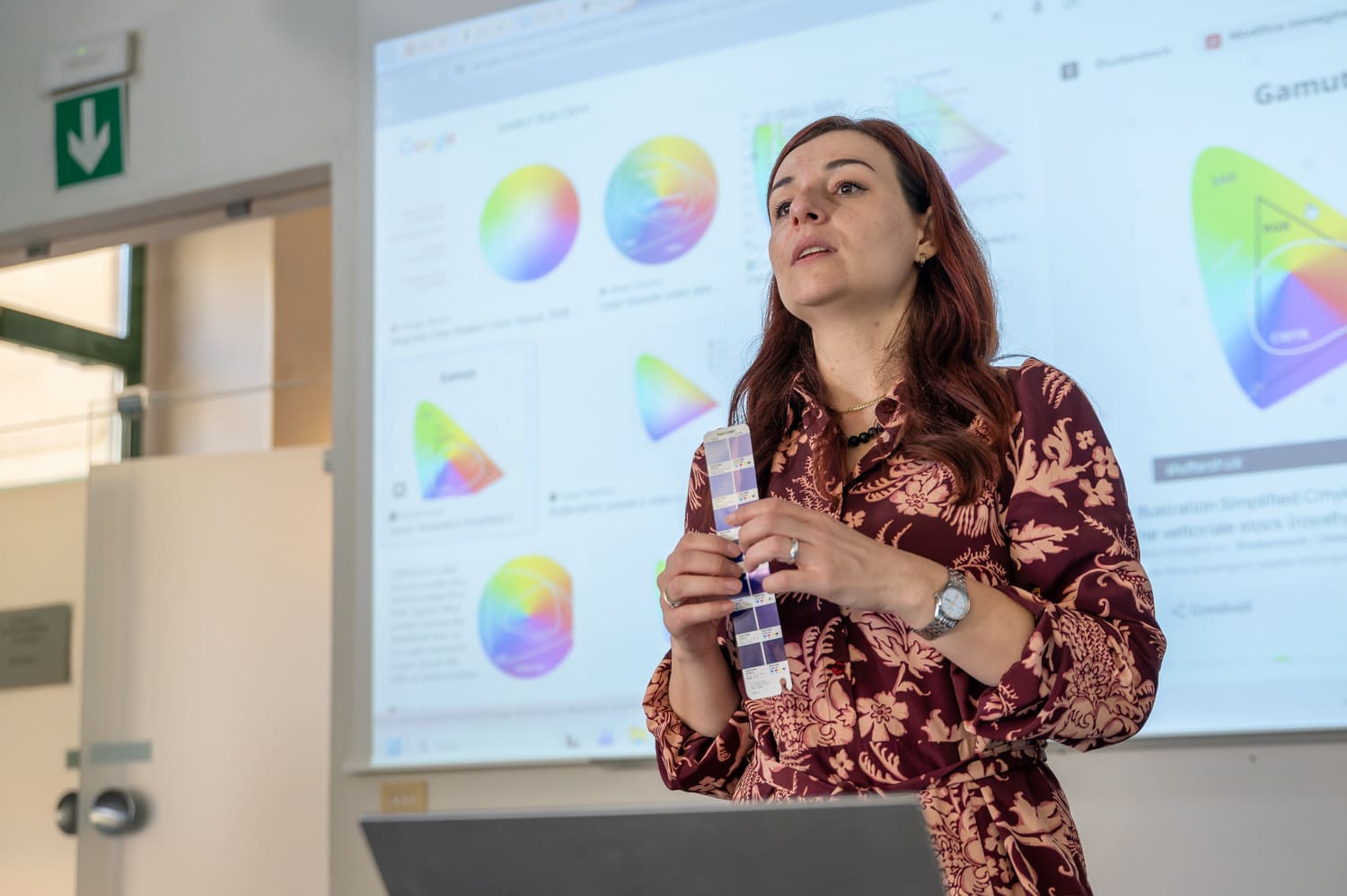 Donna tiene palette colori durante una presentazione su teoria dei colori. Schermo con grafici sullo sfondo.