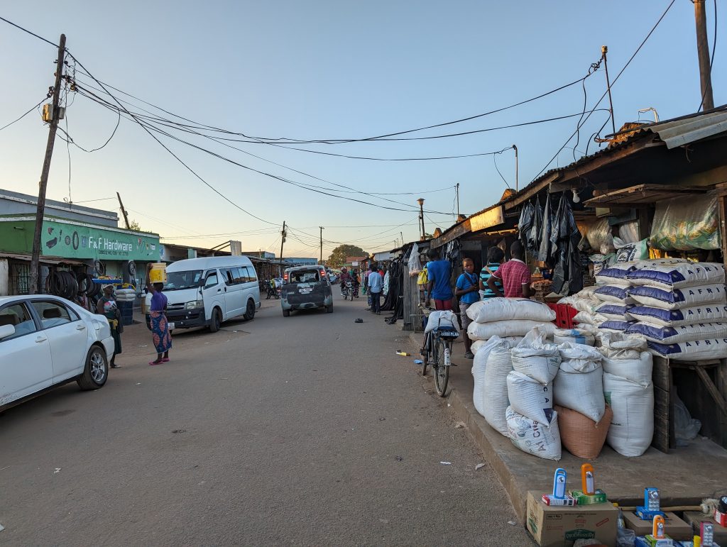 Strada con mercatino locale, automobili parcheggiate e persone che camminano accanto a sacchi e bancarelle.