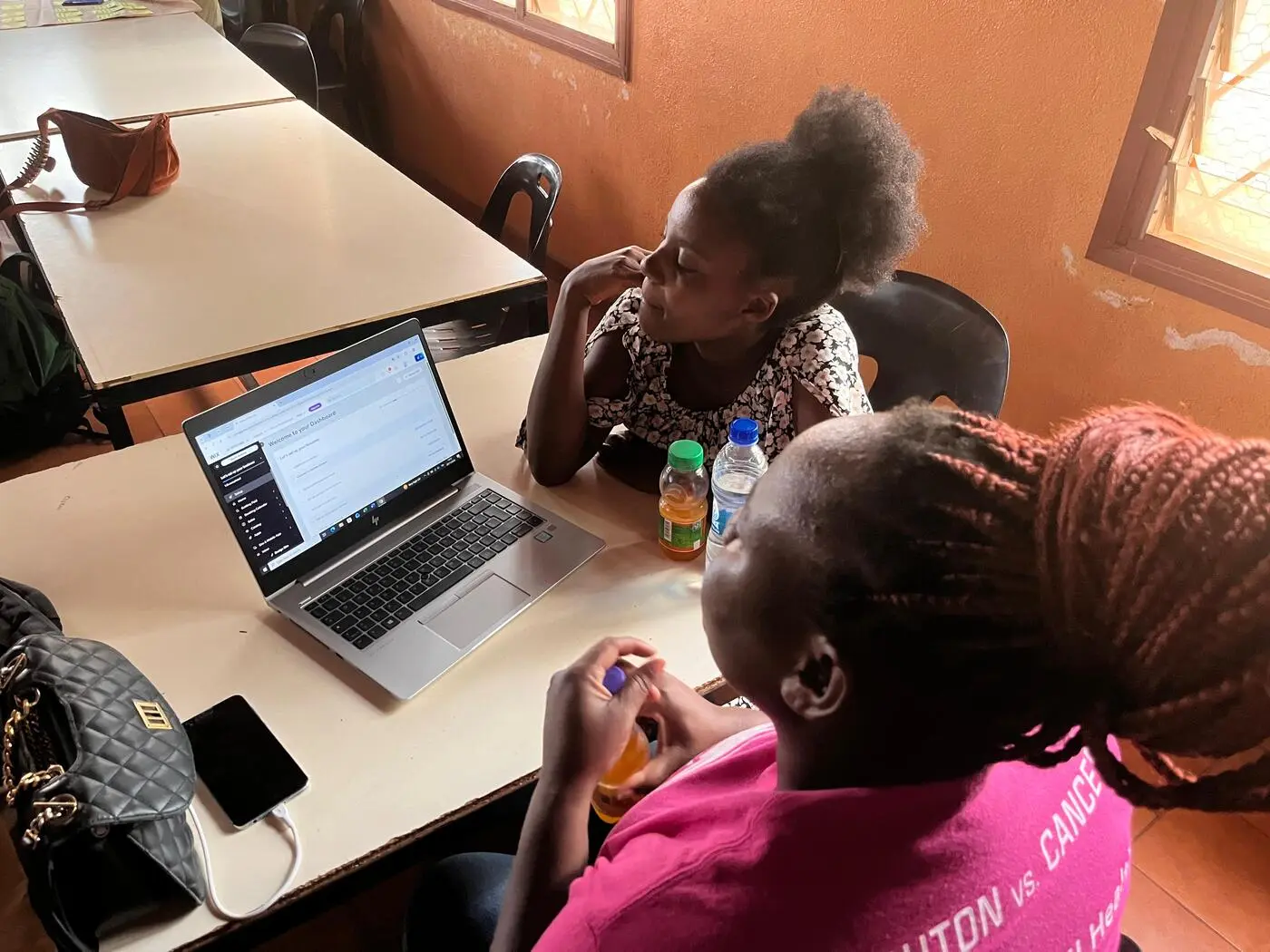 Due persone lavorano insieme al computer, bevande sul tavolo, in un'aula con pareti arancioni e finestra luminosa.