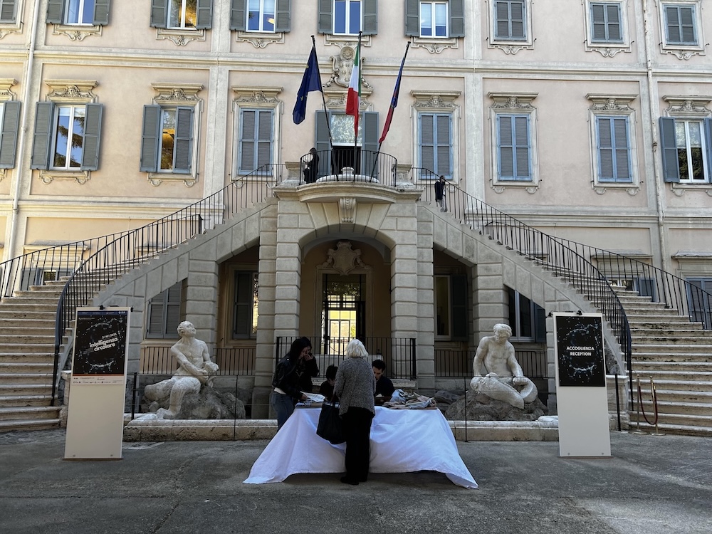 Ingresso di un elegante edificio storico con scale simmetriche e statue, persone al tavolo di benvenuto.
